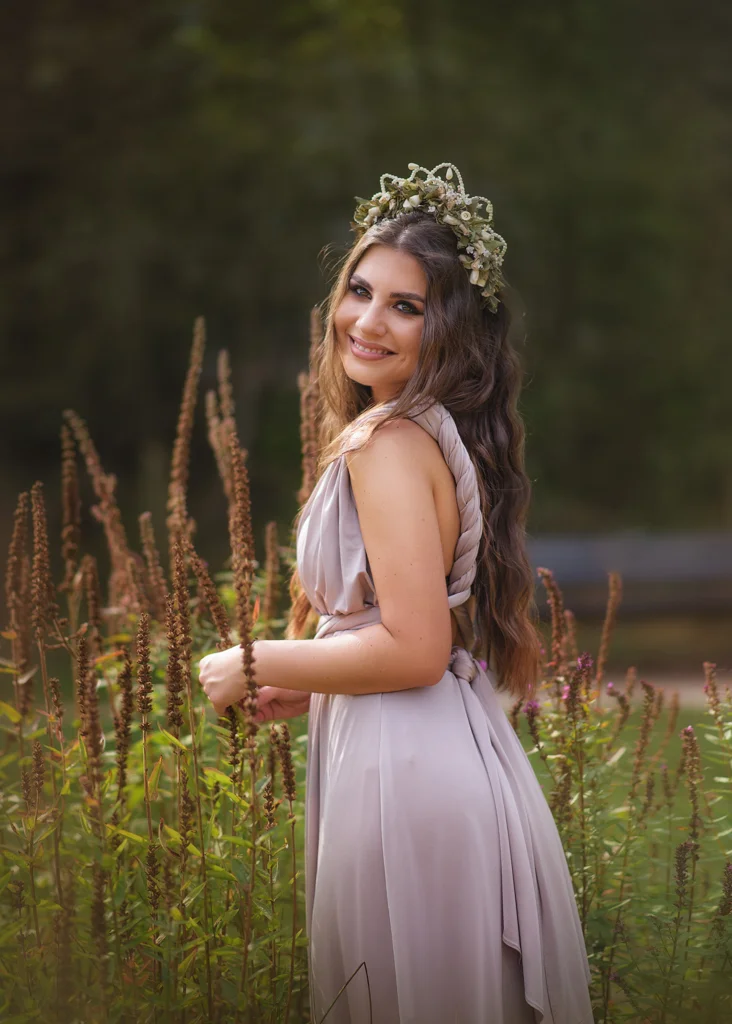 Outdoor female portrait photography of a smiling woman in a lavender dress with a floral crown standing near tall wildflowers.