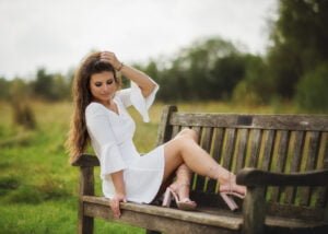 Outdoor female portrait photography with a woman in a white dress seated on a wooden bench in a grassy field.