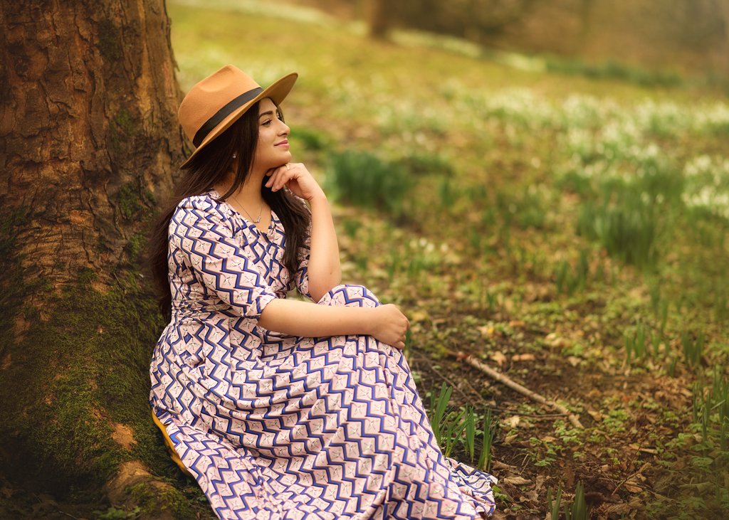 Woman in a patterned dress and hat sitting serenely by a tree in a blossoming spring field for her outdoor birthday photoshoot.