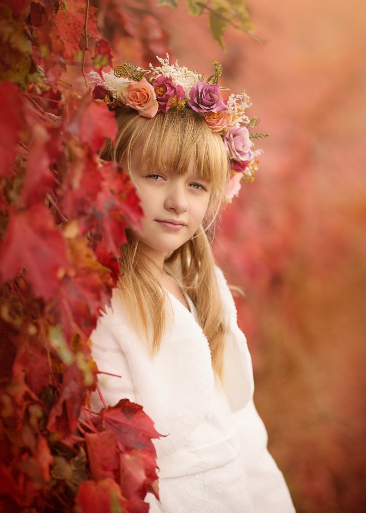 A contemplative young girl in her first holy communion attire, framed by autumn's fiery foliage, wears a crown of vibrant flowers as a symbol of her blossoming faith.
