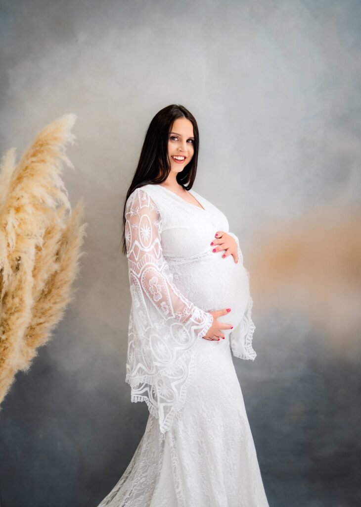 Expectant mother in a white lace dress standing beside pampas grass during a Nottingham maternity photoshoot.