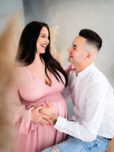 Expectant couple laughing together during a maternity photoshoot with the mother wearing a pink dress.
