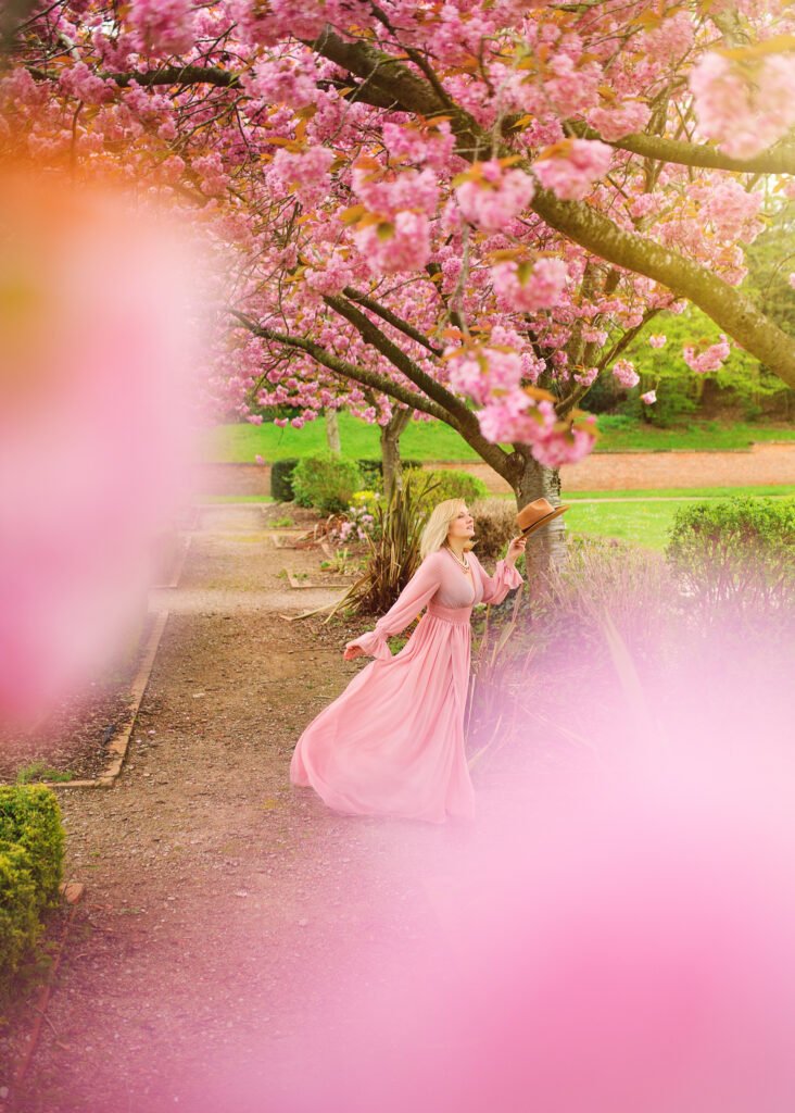 A mature woman in a dreamy pink gown reaches out to a cherry blossom tree, her gaze uplifted and joyful, in a garden path lined with vibrant spring flowers