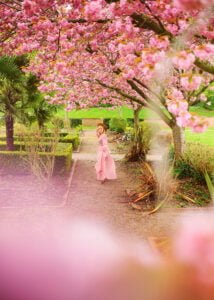 Amidst a lush springtime garden, a mature woman in a pastel pink dress looks over her shoulder with a playful smile, her presence as enchanting as the surrounding cherry blossoms.
