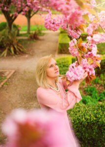 A contemplative mature woman in a soft pink dress admires cherry blossoms, her gaze filled with wonder and appreciation, amidst the verdant gardens of Nottingham.