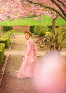 A mature woman in a pastel pink dress and stylish hat looks back with a captivating smile, strolling down a garden path framed by pink cherry blossoms in Nottingham.