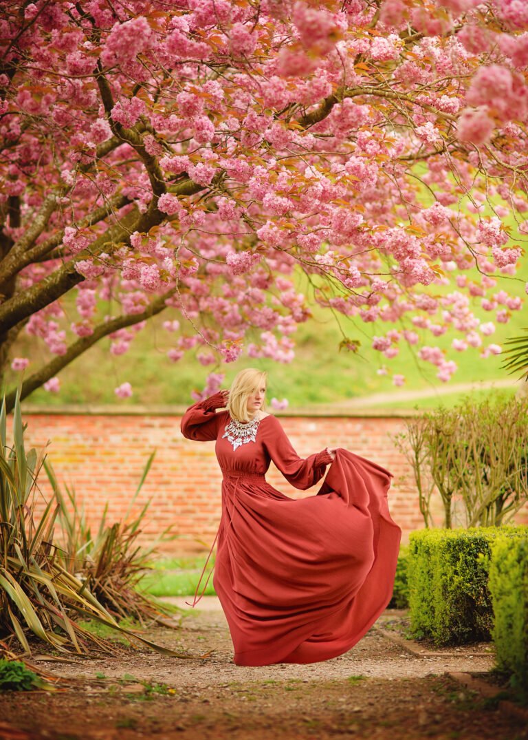 A mature woman in a flowing terracotta dress dances under a canopy of pink cherry blossoms, her movement full of life and her expression one of carefree joy.