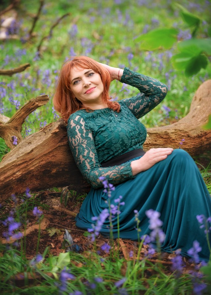 Joyful red-haired woman in a green lace dress seated by a fallen log in a bluebell wood during her outdoor birthday photoshoot.