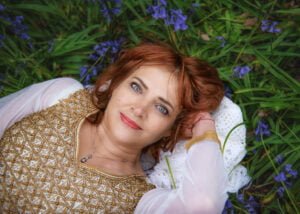 Red-haired mature woman lying gracefully among bluebells, wearing an elegant golden embroidered dress.