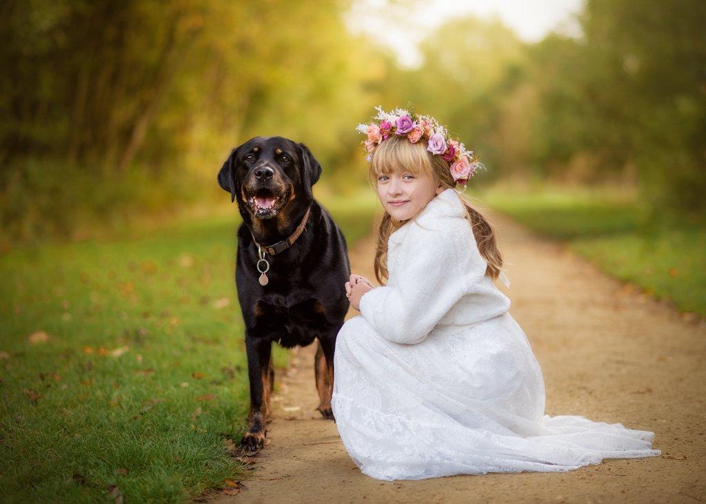 A young girl adorned with a floral crown sits beside her loyal black dog on a picturesque Nottingham trail, a testament to the timeless bond between children and their pets.