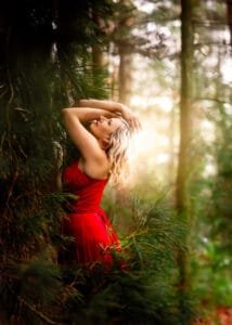 Elegant mature woman in a vibrant red dress leaning against trees with sunlight streaming through a serene woodland in Nottingham.
