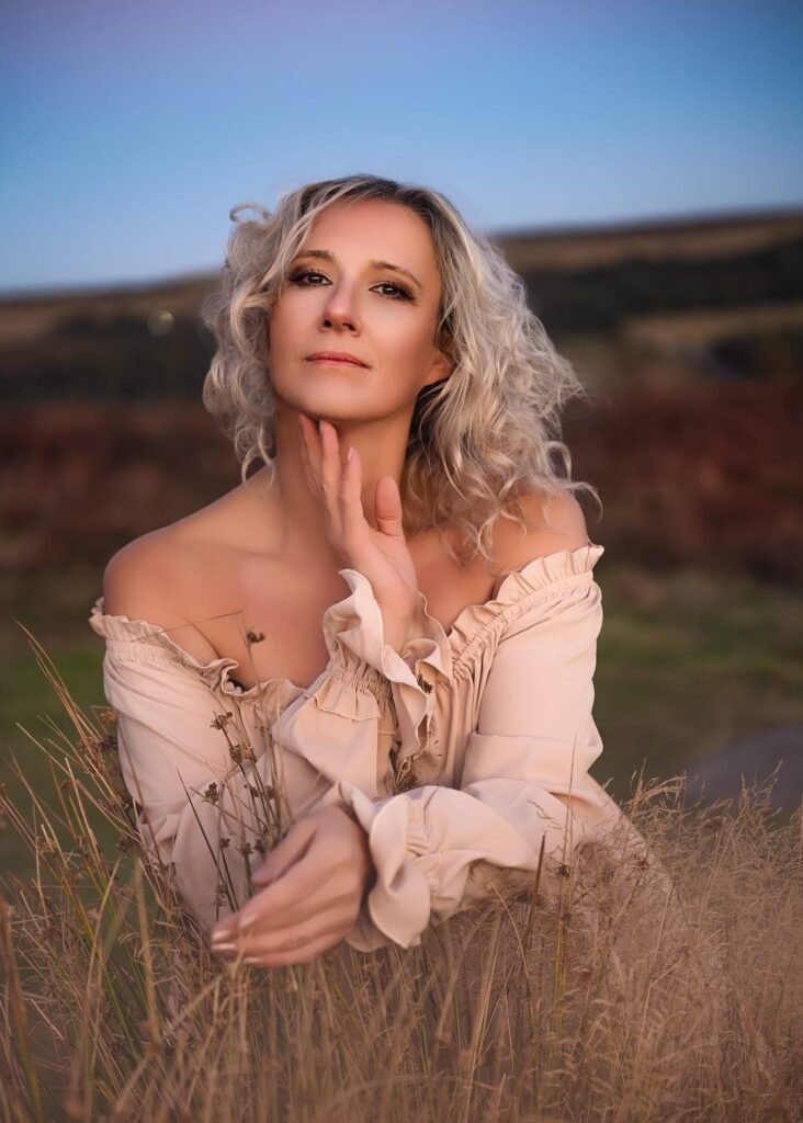 A mature blonde woman in a beige off-shoulder dress, gracefully posing in a field during golden hour.