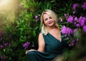 Blonde mature woman in a teal dress surrounded by purple flowers, captured in soft natural light during a portrait session in Nottingham.