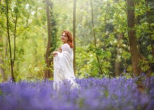 Red-haired woman in a flowing white dress walking through bluebells in a lush Nottingham woodland during a photoshoot.