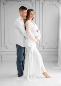 Expectant couple embracing during a maternity photoshoot with the mother wearing a white lace dress in Nottingham studio.