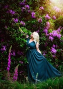 Mature woman in a flowing teal dress reaching for vibrant purple blossoms during a portrait session in Nottingham.