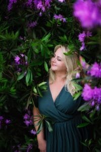 Blonde mature woman in a teal dress smiling peacefully surrounded by vibrant purple flowers in Nottingham.