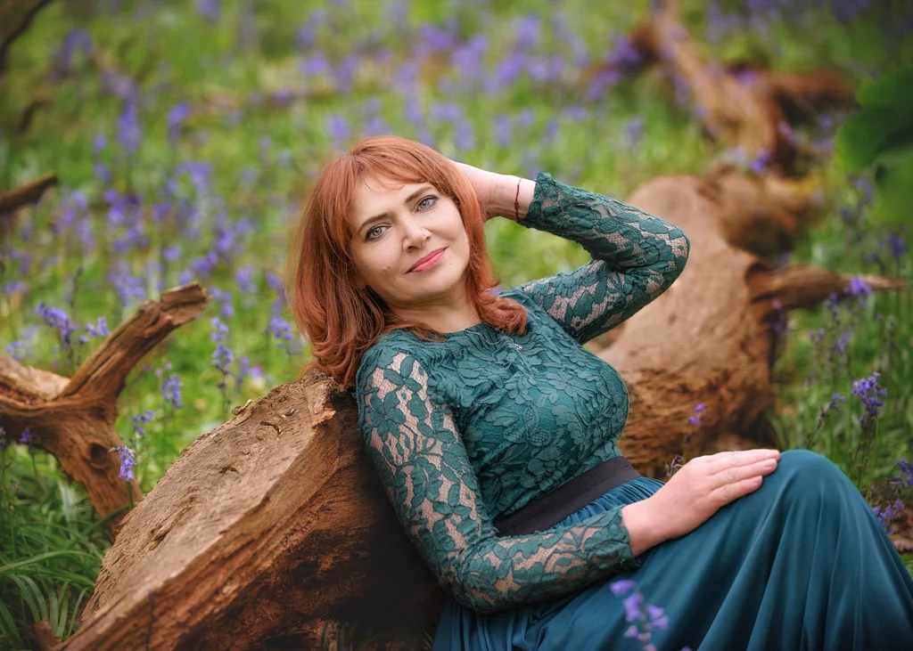 Red-haired mature woman in a lace green dress reclining on a log in Nottingham’s lush woodland.