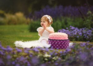 Toddler in a white dress enjoying her 1st Birthday Photoshoot outdoors with a pink cake smash surrounded by flowers.