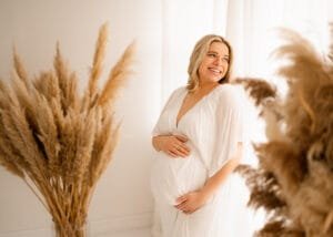Glowing mom-to-be in a white maternity gown during a baby bump photoshoot, surrounded by pampas grass in a studio.
