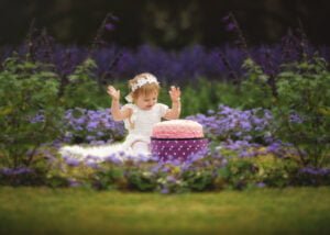 Toddler celebrating her 1st Birthday Photoshoot with a cake smash surrounded by vibrant purple flowers in a garden.