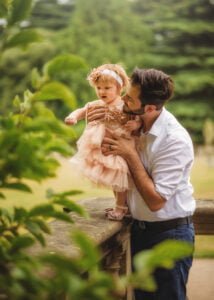 Father lovingly holding his toddler daughter during a 1st Birthday Photoshoot in a lush outdoor garden.