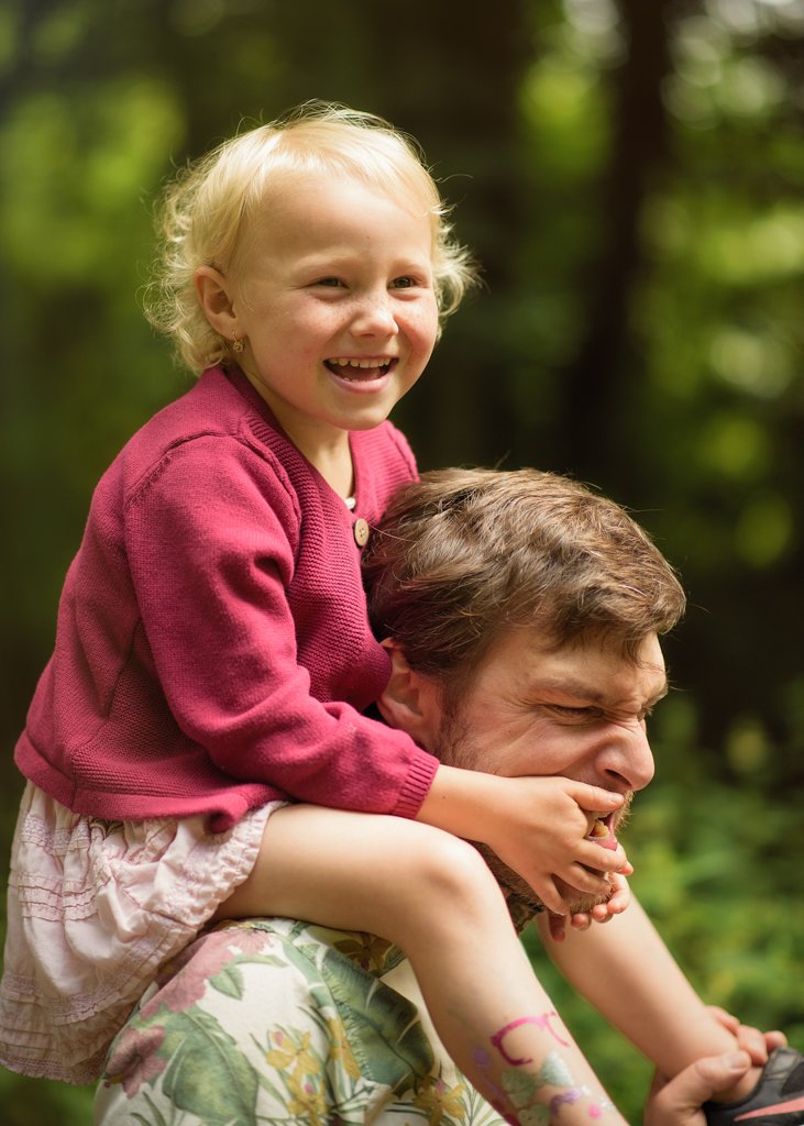 A Nottingham father smiles as he carries his joyful daughter on his shoulders, both enjoying a playful moment in the great outdoors.