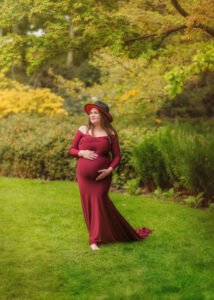 A serene pregnant woman stands in a lush garden, her elegant burgundy dress and stylish hat complementing the autumn foliage around her.