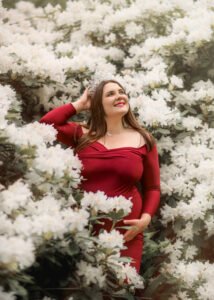 A radiant pregnant woman in a burgundy dress stands amidst a sea of white flowers, her tiara sparkling as she looks up with hopeful anticipation.