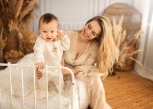 Baby standing in a crib while smiling mother watches during a baby photography session in Eastwood.