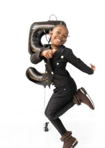 Energetic birthday photography of a young girl in a black outfit, jumping joyfully with a number 5 balloon.
