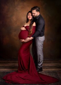Loving couple celebrates their growing family during a maternity session with the mother-to-be in a red gown and the father-to-be embracing her.