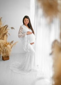 Elegant mom-to-be in a white lace dress poses near pampas grass during a serene maternity session.