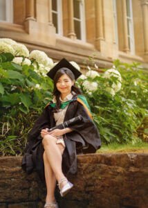 Female graduate in cap and gown sitting by flowers on Nottingham University campus
