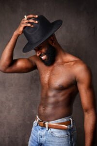 Confident, shirtless men’s photoshoot featuring a man in a black hat, smiling naturally.