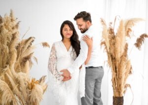 Expectant couple sharing a sweet moment during a maternity session, with the mother-to-be in a white lace dress surrounded by pampas grass.