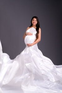 Elegant mother-to-be in a flowing white satin gown during a maternity session in a dramatic studio setting.