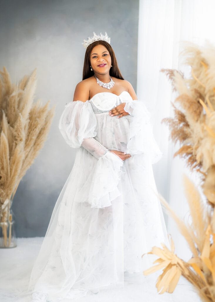 Regal mom-to-be in a white gown and crown during a maternity session surrounded by pampas grass.