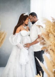 Expectant parents share a loving moment during a maternity session in a white-themed studio with pampas grass.
