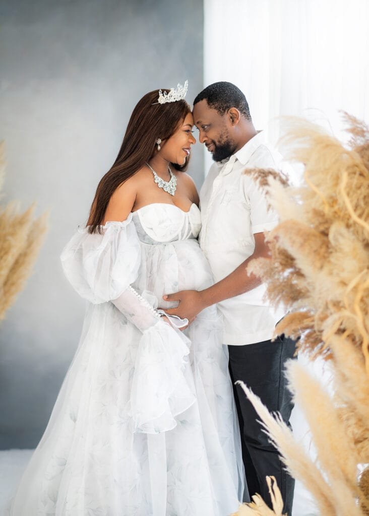 Expectant parents share a loving moment during a maternity session in a white-themed studio with pampas grass.