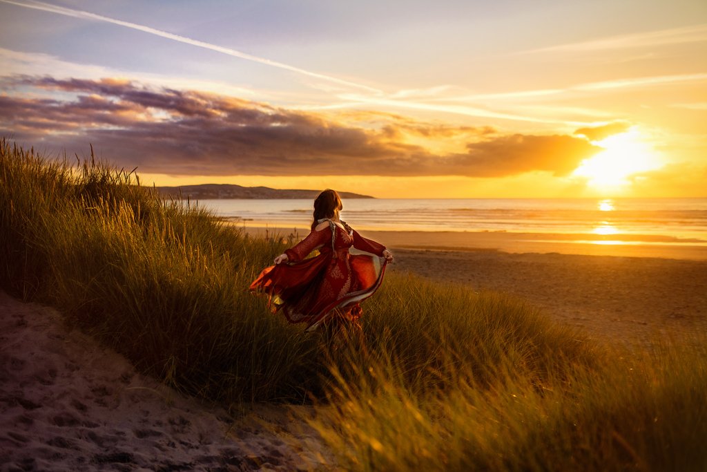 A woman in a vibrant red dress runs joyously along the beach at sunset in a heartwarming outdoor birthday photoshoot.