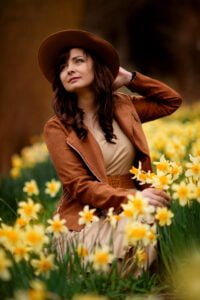 A woman with an air of thoughtful elegance adjusts her chic hat, surrounded by the vibrant yellow of daffodils, her gaze lifted in a moment of optimistic reflection