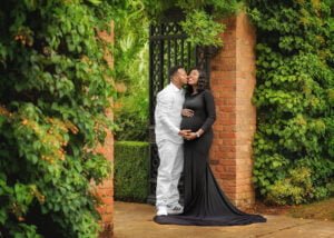 Expectant couple posing by a vine-covered gate, with the mother-to-be in a flowing black gown during a maternity photoshoot in Nottingham.