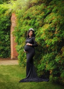 A glowing expectant queen in an elegant black gown stands proud and joyful amidst the greenery, celebrating her maternity journey.