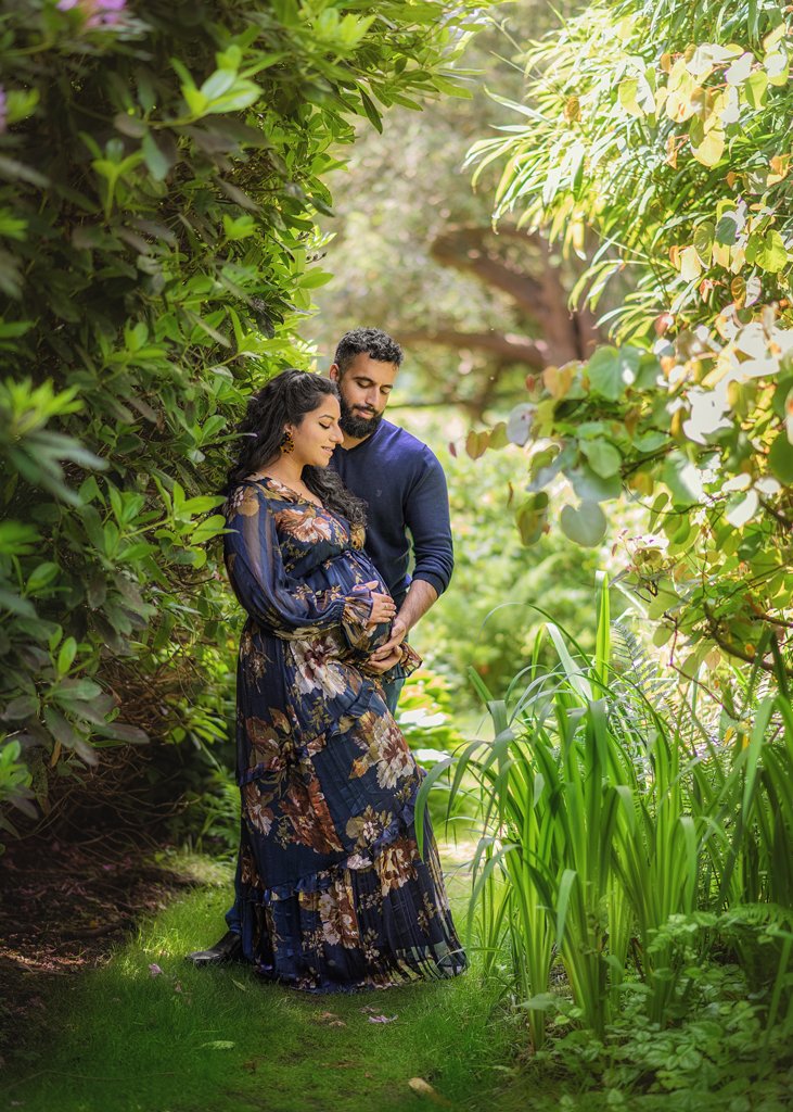 An expectant couple shares a quiet moment in a lush garden, the mother-to-be in a floral gown that echoes the surrounding greenery.