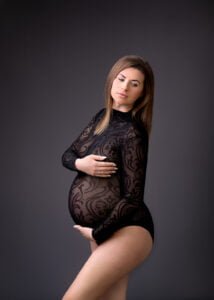 A poised mother-to-be in a patterned bodysuit exudes confidence and beauty against a soft grey backdrop, embracing her baby bump with gentle hands.