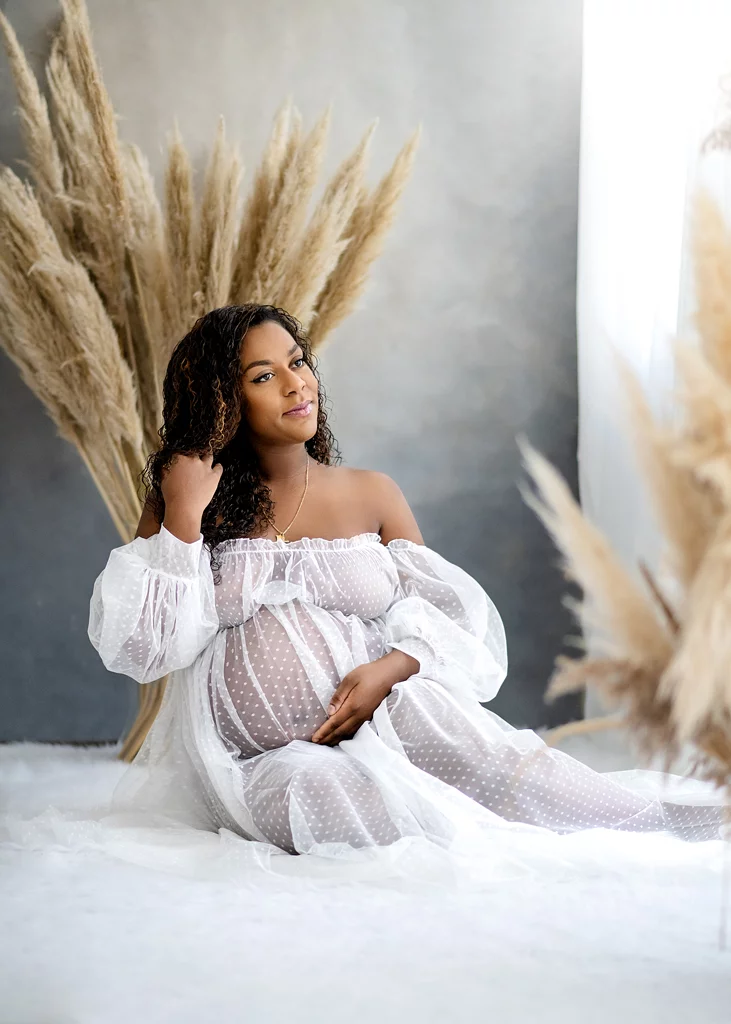 Radiant expectant mother in a sheer polka-dot maternity gown sitting gracefully near pampas grass during her maternity session.