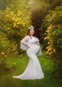 A beaming expectant mother adorned with a tiara poses elegantly in a white lace gown, surrounded by the vibrant yellow of spring flowers in a lush garden.