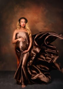 A majestic portrait of an expectant mother draped in a luxurious bronze satin gown, her hand placed gently over her baby bump, against a dramatic, mottled backdrop