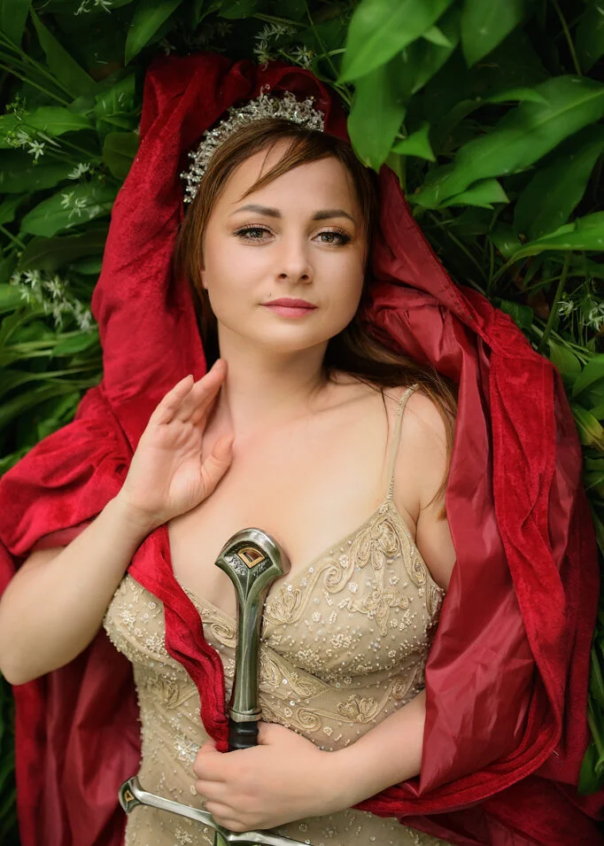 Enthroned in greenery, a woman in a regal red cloak and tiara wields a sword at her outdoor birthday photoshoot.